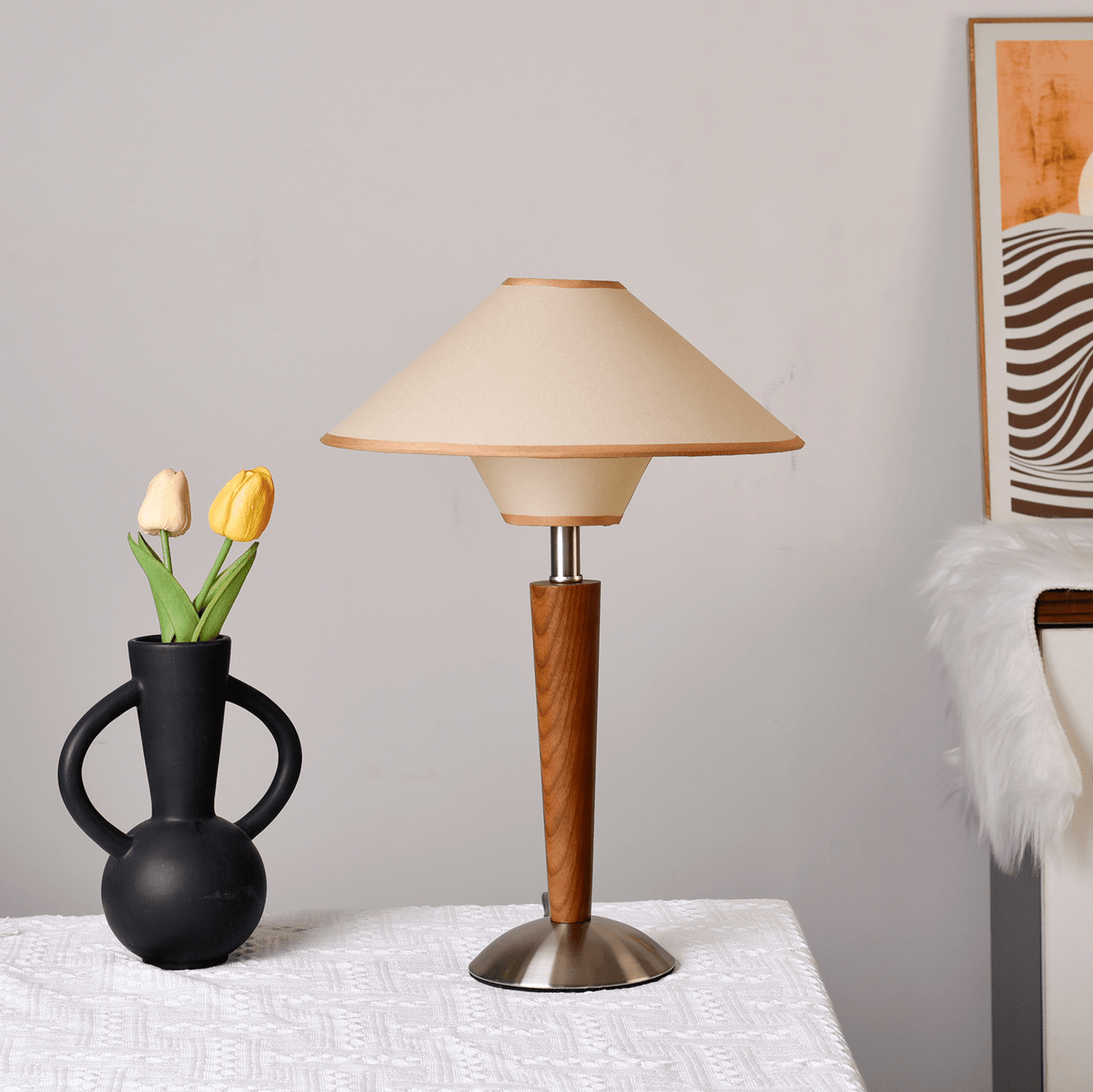 Table lamp with wooden base and beige shade next to a black vase with yellow tulips on a white surface.