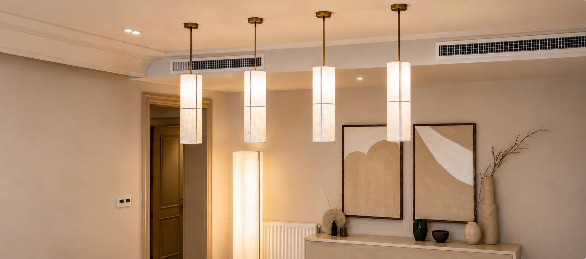 Four modern cylindrical pendant lights with white textured shades and gold-toned rods hanging over a console table in a warm, contemporary interior.